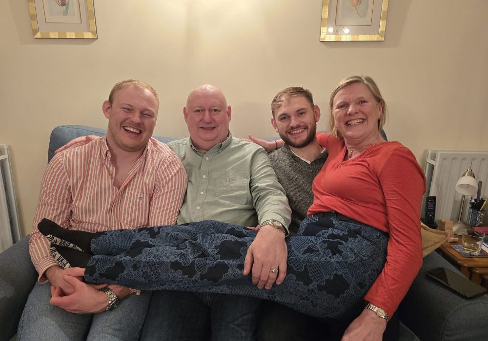 Three males sat next to each other on the sofa, mum is sat with her legs across their knees