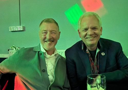 Picture of two men, sat next to each other. They look like they're on an evening out and the lighting is green.