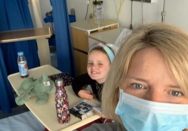 Mother wearing a face mask, standing next to hospital bed that daughter is in