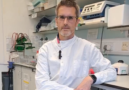 Colin, in his lab, sat next to his research bench wearing his white lab coat.