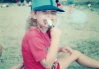 Heather, wearing a baseball cap and red tshirt, licking an ace cream, sitting on the beach