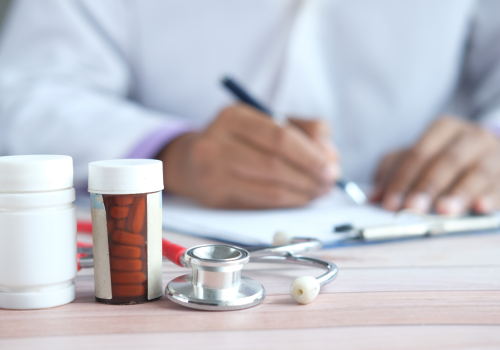 Hands writing on paper on a desk in the background. With a bottle of tablets and a stethoscope in the foreground.